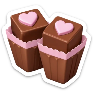 Two square milk chocolate pralines, Each praline is a sharp-edged cube with smooth glossy chocolate coating, placed in a small white fluted paper candy cup. On top of each cube is a flat pastel pink heart-shaped icing sticker