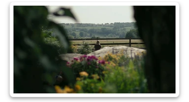"Two shot" of a person in the foreground, blurred plants in the foreground (frame within a frame), a wooden fence and colorfull flowers in the midground, Poland, rolling hills in the background, cinematic depth of field, layered composition, natural lighting sticker