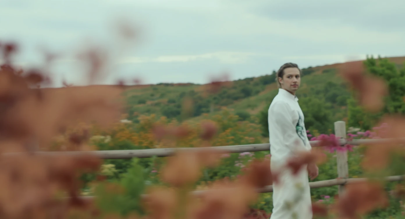 A portrait of a white person in the foreground, blurred plants in the foreground, a wooden fence and colorfull flowers in the midground, rolling hills in the background, cinematic depth of field, layered composition, natural lighting emoji