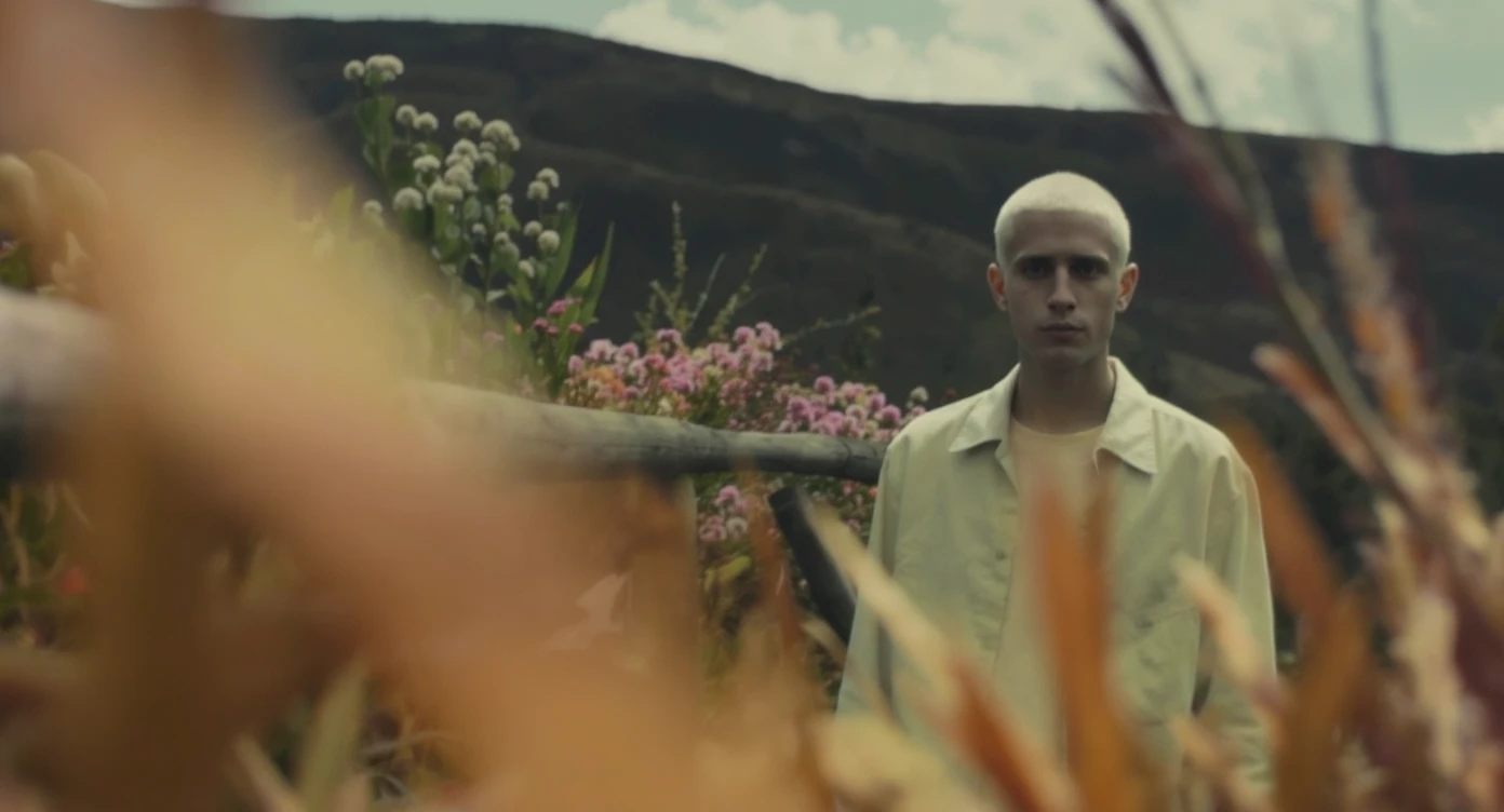 A portrait of a white person in the foreground, blurred plants in the foreground, a wooden fence and colorfull flowers in the midground, rolling hills in the background, cinematic depth of field, layered composition, natural lighting emoji