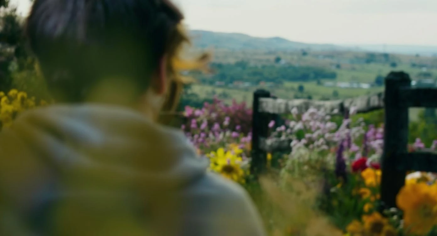 A portrait of a person in the foreground, blurred plants in the foreground (frame within a frame), a wooden fence and colorfull flowers in the midground, rolling hills in the background, cinematic depth of field, layered composition, natural lighting emoji