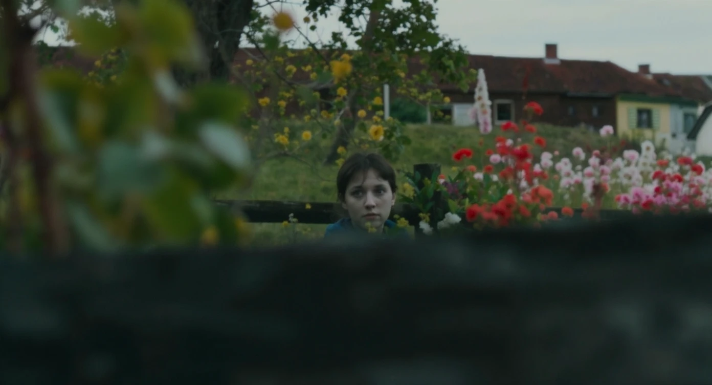 A portrait of a person in the foreground, blurred plants in the foreground (frame within a frame), a wooden fence and colorfull flowers in the midground, Poland, rolling hills in the background, cinematic depth of field, layered composition, natural lighting emoji