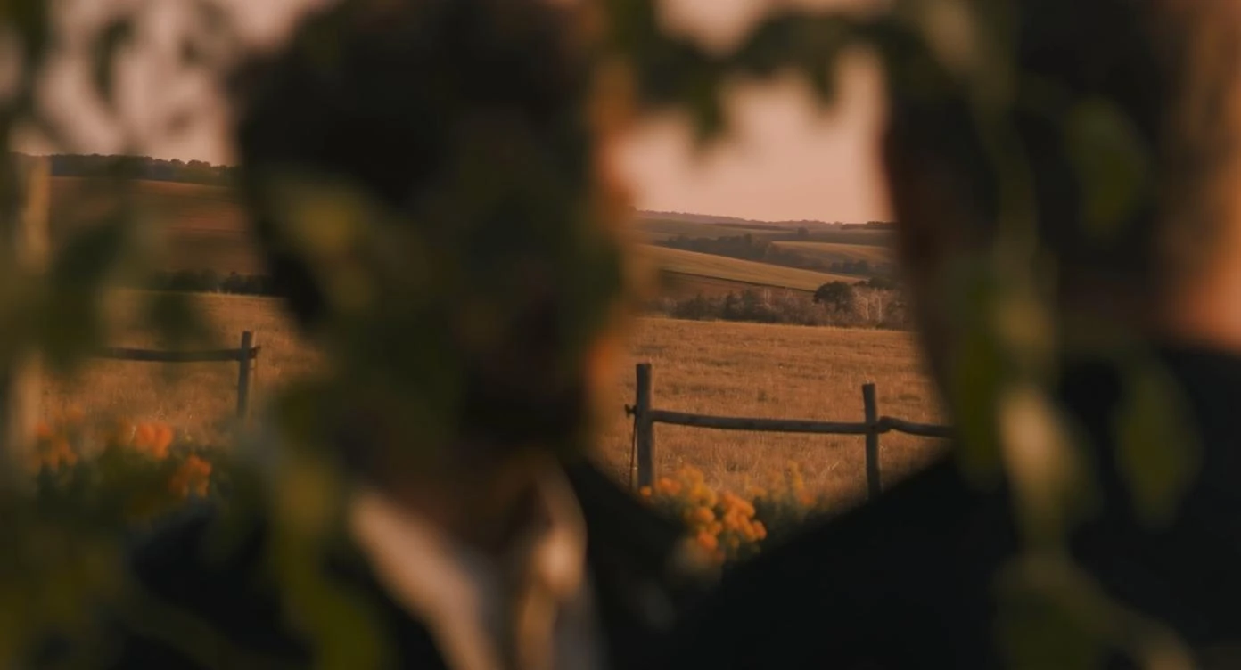 "Two shot" in the foreground, blurred plants in the foreground (frame within a frame), a wooden fence and colorfull flowers in the midground, Poland, rolling hills in the background, cinematic depth of field, layered composition, natural lighting emoji