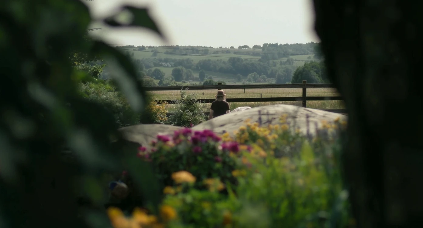 "Two shot" of a person in the foreground, blurred plants in the foreground (frame within a frame), a wooden fence and colorfull flowers in the midground, Poland, rolling hills in the background, cinematic depth of field, layered composition, natural lighting emoji