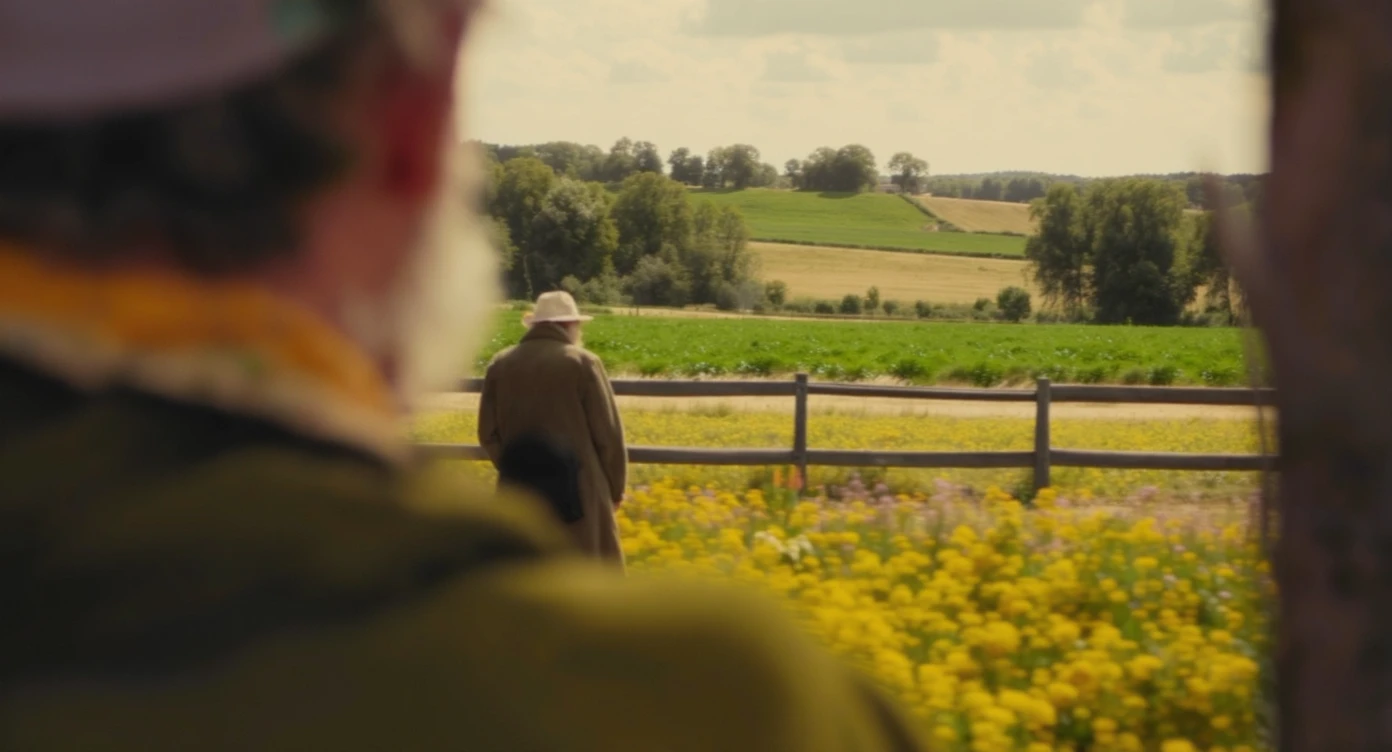 "Two shot" of a person in the foreground, blurred plants in the foreground (frame within a frame), a wooden fence and colorfull flowers in the midground, Poland, rolling hills in the background, cinematic depth of field, layered composition, natural lighting emoji